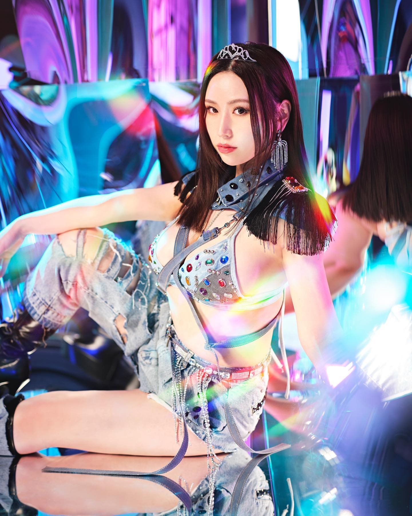 Ai Sayama como L.V com figurino prateado e reflexos coloridos